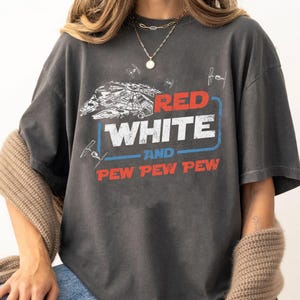 Puede incluir: Una camiseta gris oscura con un gráfico de estilo vintage que presenta una nave espacial, las palabras "Red White and Pew Pew Pew" y acentos rojos, blancos y azules.