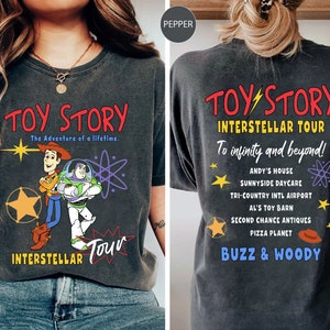 May include: Dark gray t-shirt featuring the words "TOY STORY" in red, with Woody and Buzz Lightyear graphics. The front of the shirt also says "INTERSTELLAR TOUR". The back of the shirt says "INTERSTELLAR TOUR" and "To infinity and beyond!" with a list of locations.