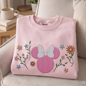 Pu&ograve; includere: Una felpa girocollo rosa chiaro con una silhouette ricamata di Minnie Mouse rosa con un fiocco bianco al centro, circondata da vari piccoli fiori di campo ricamati nelle tonalit&agrave; del blu, dell'arancione e del marrone sul davanti.