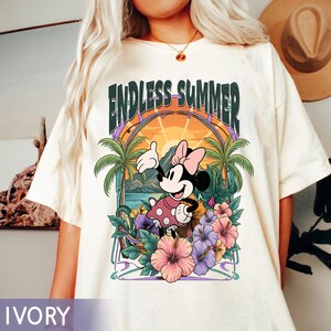 May include: Ivory-colored t-shirt featuring a graphic of Minnie Mouse in a tropical setting. The design includes the text "ENDLESS SUMMER" above Minnie, palm trees, flowers, and a sunset. The shirt is a relaxed fit.