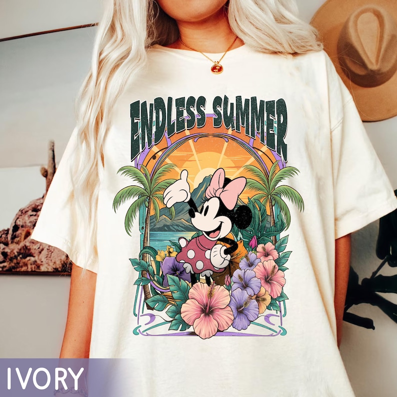 May include: Ivory-colored t-shirt featuring a graphic of Minnie Mouse in a tropical setting. The design includes the text "ENDLESS SUMMER" above Minnie, palm trees, flowers, and a sunset. The shirt is a relaxed fit.