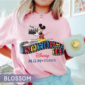 Pu&ograve; includere: T-shirt rosa chiaro con una grafica di Mickey Mouse che tiene una clapperboard, in piedi su una bobina di film. &Egrave; visibile anche il logo Disney MGM Studios. Nella foto &egrave; presente una tazza con una faccina sorridente.