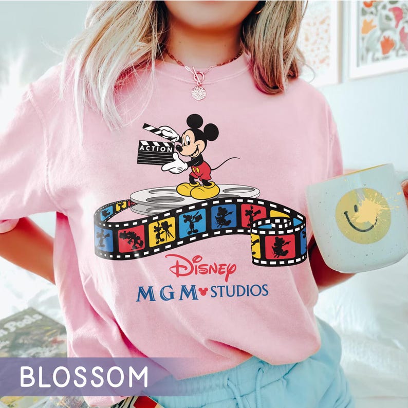 Pu&ograve; includere: T-shirt rosa chiaro con una grafica di Mickey Mouse che tiene una clapperboard, in piedi su una bobina di film. &Egrave; visibile anche il logo Disney MGM Studios. Nella foto &egrave; presente una tazza con una faccina sorridente.