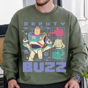 May include: Olive green sweatshirt featuring a graphic of Buzz Lightyear from Pixar's Toy Story. The design includes the words "DEPUTY BUZZ" in a purple, blocky font, and several images of Buzz in different poses.