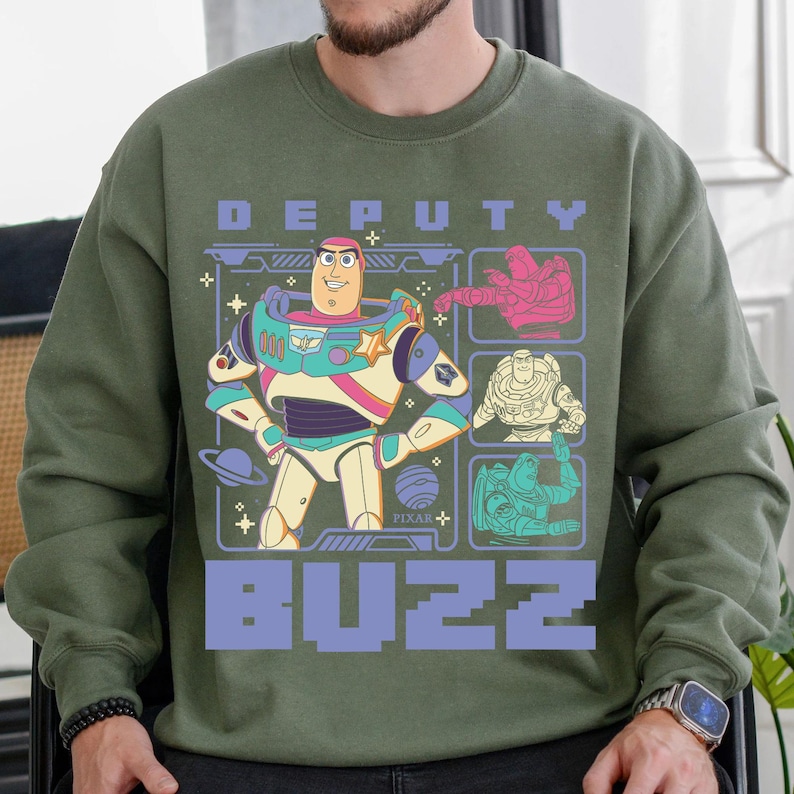 May include: Olive green sweatshirt featuring a graphic of Buzz Lightyear from Pixar's Toy Story. The design includes the words "DEPUTY BUZZ" in a purple, blocky font, and several images of Buzz in different poses.
