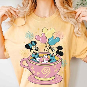 May include: Yellow t-shirt with a cartoon design of Mickey and Minnie Mouse in a pink teacup, holding hands. Balloons and fireworks are in the background. The design is playful and colourful, ideal for Disney fans.