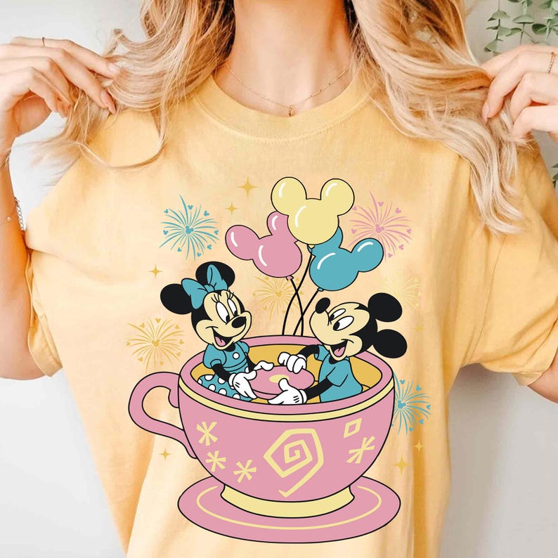 May include: Yellow t-shirt with a cartoon design of Mickey and Minnie Mouse in a pink teacup, holding hands. Balloons and fireworks are in the background. The design is playful and colourful, ideal for Disney fans.