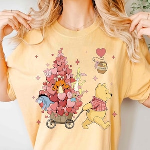 May include: Yellow t-shirt with a graphic of Winnie the Pooh pulling a cart filled with hearts and Disney characters. A jar of "Hunny" and pink hearts are also in the design. The shirt has a crew neck and short sleeves.