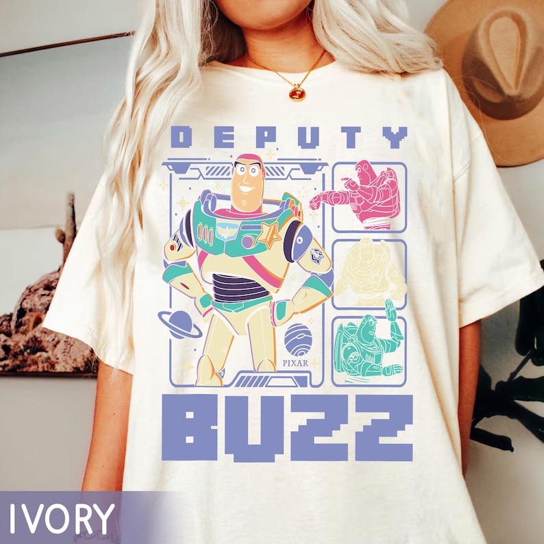 May include: Ivory-colored t-shirt featuring a graphic of Buzz Lightyear from the movie Toy Story. The design includes the words "DEPUTY" and "BUZZ" in a blue font, along with illustrations of Buzz in different poses. The Pixar logo is also visible.