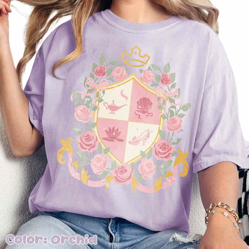 Puede incluir: Camiseta color orqu&iacute;dea con un dise&ntilde;o de escudo que presenta una corona, rosas e iconos de cuentos de hadas. El escudo incluye una l&aacute;mpara de genio, una concha, una flor de loto y un zapato de cristal. La camiseta tiene un corte holgado.