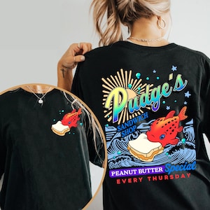May include: Black t-shirt featuring a colorful graphic of a red fish biting a peanut butter sandwich. The back of the shirt has the text "Pudge's Sandwich Shop" and "Peanut Butter Special Every Thursday".