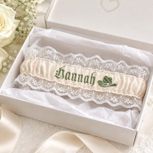 May include: A cream-colored satin garter featuring white lace trim on the top and bottom edges. The center is embroidered with the name Hannah in dark green gothic-style lettering, accompanied by a small green cowboy hat icon with a white heart.