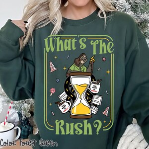 Puede incluir: Sudadera verde bosque con un gr&aacute;fico que presenta un perezoso, un reloj de arena y el texto "What's The Rush?". El dise&ntilde;o incluye tazas de caf&eacute; y conos de tr&aacute;fico. El texto "You want it when?" est&aacute; en las tazas de caf&eacute;.