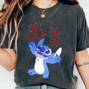 May include: Dark gray t-shirt featuring a blue cartoon character with white accents, celebrating with red and white fireworks. The character has large eyes and a wide smile. The shirt is a casual style.