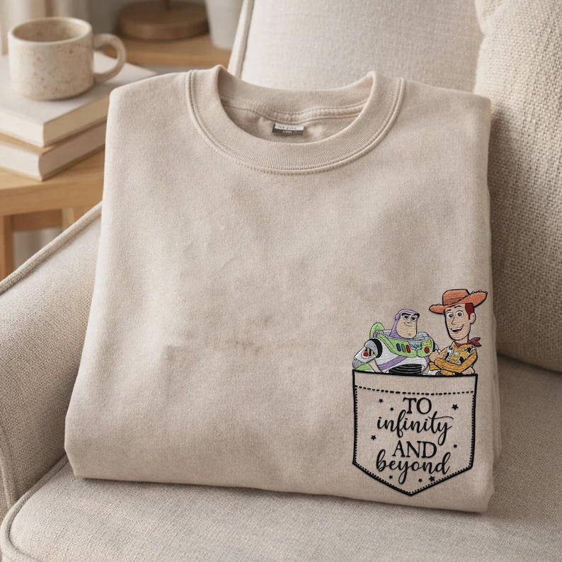 May include: A beige crewneck sweatshirt featuring a graphic of Buzz Lightyear and Woody from Toy Story appearing to peek out of a pocket. Below the characters, the text reads To infinity AND beyond in a black script font on the sweatshirt.
