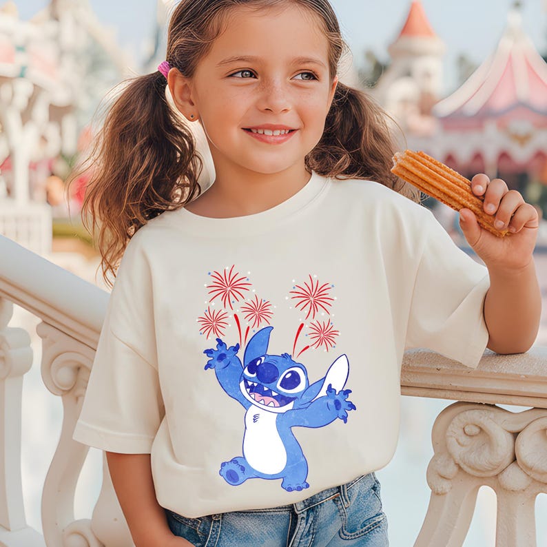 May include: A cream-colored t-shirt featuring a blue cartoon character with red and white fireworks. The character is smiling with its arms outstretched. The shirt is worn by a young girl holding a churro.