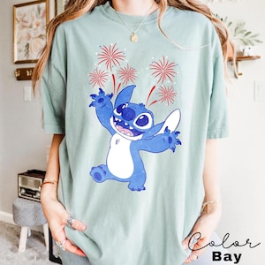 May include: A light green t-shirt featuring a blue cartoon character with white accents, arms outstretched, and a smiling expression. Red and white fireworks are above the character. The text "Color Bay" is in the bottom right corner.