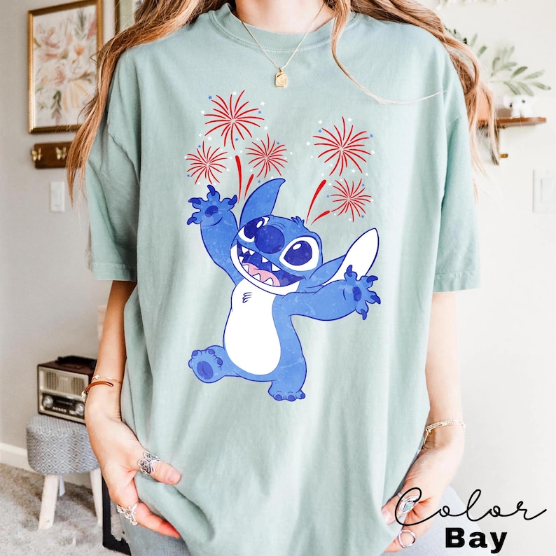 May include: A light green t-shirt featuring a blue cartoon character with white accents, arms outstretched, and a smiling expression. Red and white fireworks are above the character. The text "Color Bay" is in the bottom right corner.