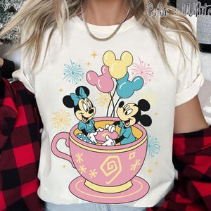 May include: White t-shirt with a colourful graphic of Mickey and Minnie Mouse in a pink teacup with balloons. The design includes fireworks and the text "Color: White". The shirt has short sleeves and a round neckline.