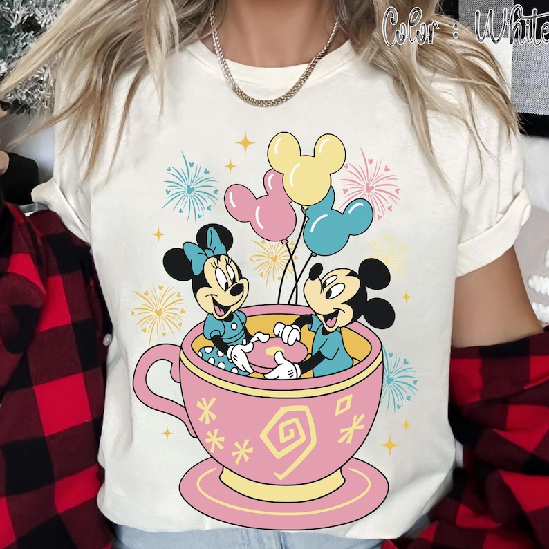 May include: White t-shirt with a colourful graphic of Mickey and Minnie Mouse in a pink teacup with balloons. The design includes fireworks and the text "Color: White". The shirt has short sleeves and a round neckline.