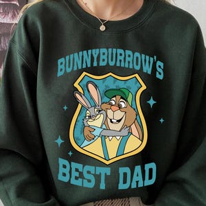 Puede incluir: Sudadera verde oscuro con un gr&aacute;fico de dibujos animados de dos conejos abraz&aacute;ndose, con el texto "Bunnyburrow's Best Dad" en color turquesa. El gr&aacute;fico est&aacute; dentro de una forma de escudo, con estrellas turquesas. Se ve un collar dorado.