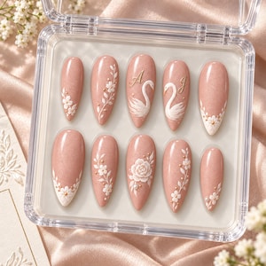 May include: A set of ten almond-shaped press-on nails in a shimmering dusty rose finish, displayed in a clear plastic case. The nails feature white 3D floral accents, delicate vine patterns, and two nails decorated with white swan motifs and gold letters A and J.