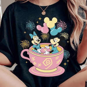 May include: Black t-shirt with a vibrant graphic of Mickey and Minnie Mouse in a pink teacup, surrounded by fireworks and balloons. The design incorporates yellow, pink, and blue, offering a cheerful and celebratory aesthetic.