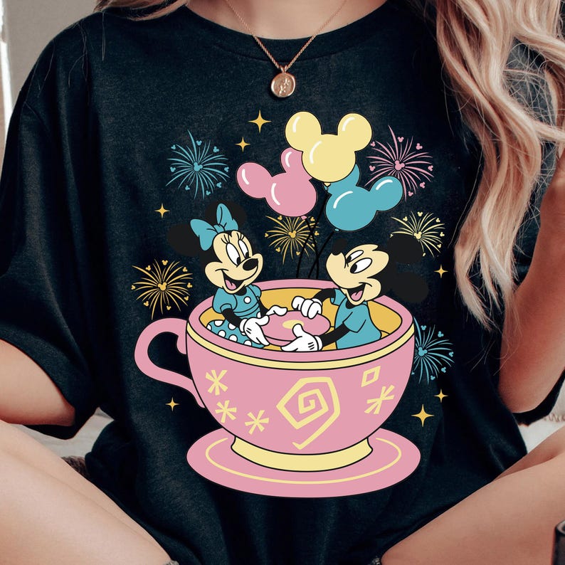 May include: Black t-shirt with a vibrant graphic of Mickey and Minnie Mouse in a pink teacup, surrounded by fireworks and balloons. The design incorporates yellow, pink, and blue, offering a cheerful and celebratory aesthetic.