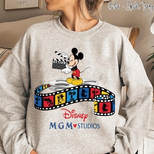 Pu&ograve; includere: Felpa grigio sportivo con grafica di Topolino che tiene una clapperboard con la scritta "ACTION". Il design include una bobina di film e una striscia di film colorata con il testo Disney MGM Studios.