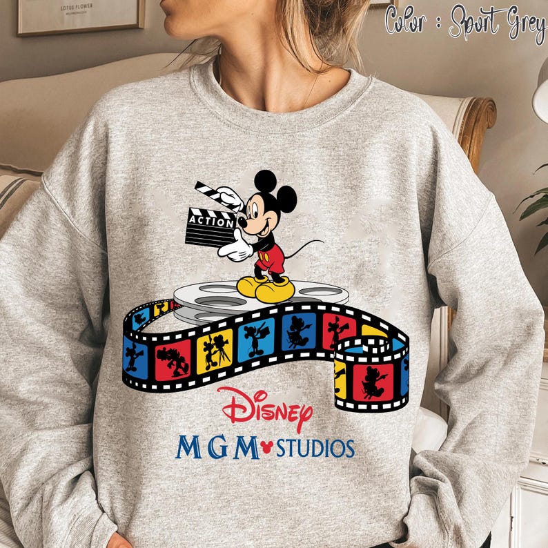 Pu&ograve; includere: Felpa grigio sportivo con grafica di Topolino che tiene una clapperboard con la scritta "ACTION". Il design include una bobina di film e una striscia di film colorata con il testo Disney MGM Studios.