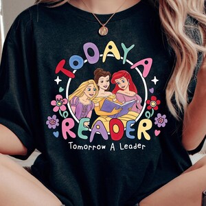 May include: Black t-shirt featuring a colorful graphic with the words "TODAY READER Tomorrow A Leader." The design includes illustrations of three princesses reading a book, surrounded by flowers and stars. The text is in a playful, colorful font.