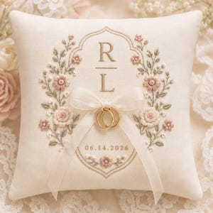 May include: A square off-white ring pillow featuring intricate embroidery of pink flowers and green leaves surrounding the letters R and L. A sheer white ribbon is tied in a bow, holding two gold rings, with the date 06.14.2026 stitched below.
