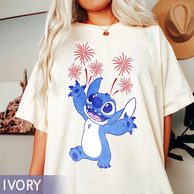 May include: Ivory-colored t-shirt featuring a blue cartoon character with a wide grin and outstretched arms, with red and white fireworks above. The word "IVORY" is printed at the bottom of the shirt.