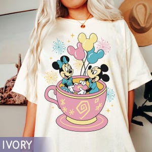 May include: Ivory t-shirt with a colourful graphic of Mickey and Minnie Mouse in a pink teacup, holding balloons. The design includes fireworks and decorative elements, creating a playful and cheerful aesthetic.