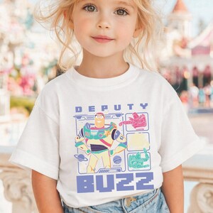 May include: White t-shirt featuring a colorful graphic of Buzz Lightyear with the text "DEPUTY BUZZ". The design includes images of Buzz Lightyear, a planet, and other space-themed elements. The shirt is worn by a young child.