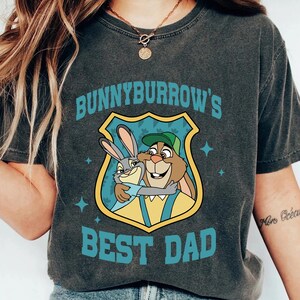 Puede incluir: Camiseta gris oscuro con un gr&aacute;fico de dibujos animados de dos conejos abraz&aacute;ndose dentro de un escudo. El texto "Bunnyburrow's Best Dad" est&aacute; impreso arriba y abajo del gr&aacute;fico en color turquesa.