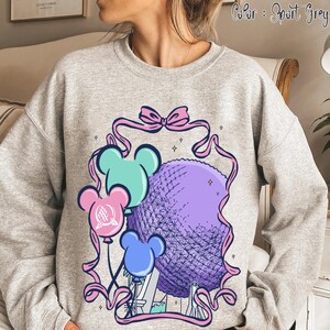 May include: Sport grey sweatshirt featuring a colorful graphic. The design includes a purple sphere, reminiscent of a geodesic dome, with three mouse-shaped balloons in blue, pink, and teal. Pink ribbons and a bow frame the central image.