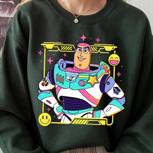 May include: Dark green sweatshirt featuring a colorful graphic of a cartoon space ranger character. The character is in a white, teal, pink, and purple space suit with a yellow star and a smiling face. The design is framed by a yellow border with smiley faces and stars.