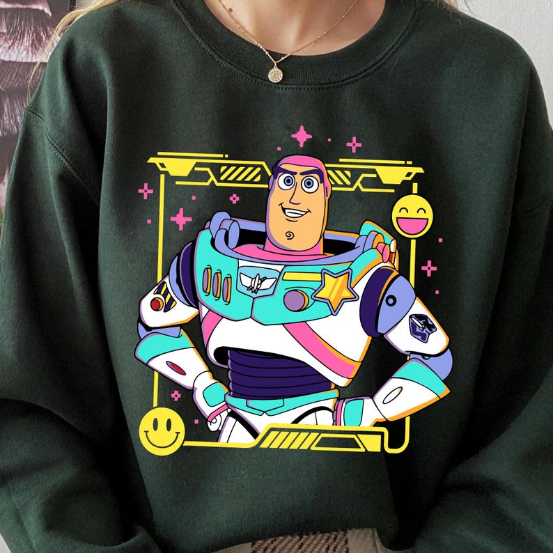 May include: Dark green sweatshirt featuring a colorful graphic of a cartoon space ranger character. The character is in a white, teal, pink, and purple space suit with a yellow star and a smiling face. The design is framed by a yellow border with smiley faces and stars.