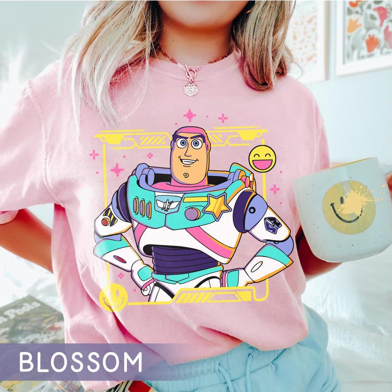 May include: A light pink t-shirt featuring a colorful graphic of Buzz Lightyear from Toy Story. The design includes a smiling Buzz, a yellow frame, and a small smiley face. The word "BLOSSOM" is printed at the bottom of the shirt.