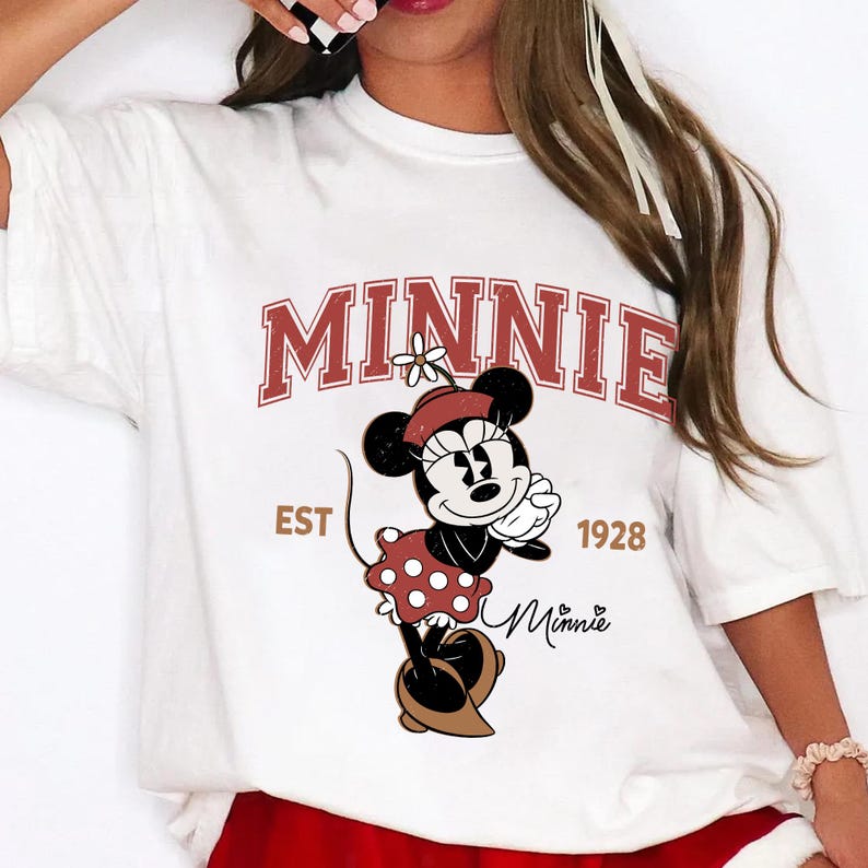 May include: White t-shirt featuring a graphic of Minnie Mouse in a red polka dot dress and a matching hat. The word "MINNIE" is printed in a maroon collegiate font above the character, with "EST 1928" below. A Minnie Mouse signature is also present.
