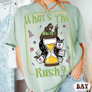 Puede incluir: Camiseta verde claro con la frase "What's The Rush?" y un dise&ntilde;o de perezoso. El dise&ntilde;o incluye un reloj de arena, tazas de caf&eacute; y elementos de carretera. La camiseta tiene cuello redondo y mangas cortas.