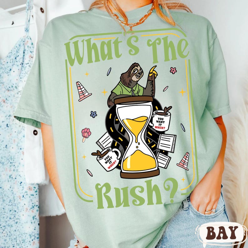 Puede incluir: Camiseta verde claro con la frase "What's The Rush?" y un dise&ntilde;o de perezoso. El dise&ntilde;o incluye un reloj de arena, tazas de caf&eacute; y elementos de carretera. La camiseta tiene cuello redondo y mangas cortas.