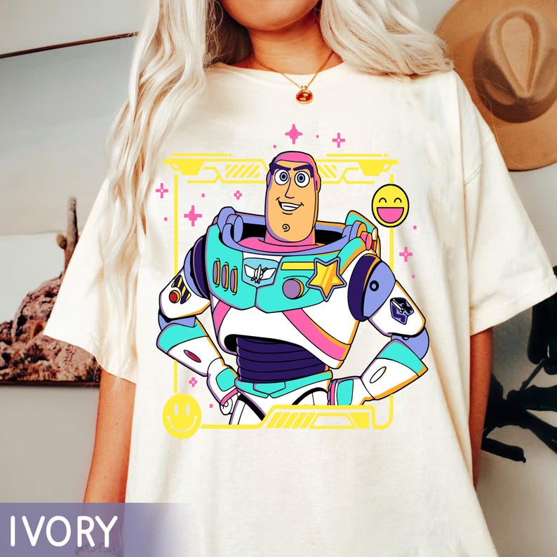 May include: Ivory-colored t-shirt featuring a colorful graphic of Buzz Lightyear from Toy Story. The design includes a yellow frame, pink accents, and a smiling face. The shirt is a relaxed fit.
