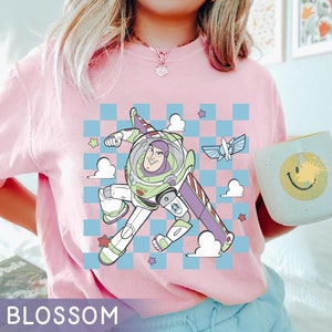 May include: Pink t-shirt with a cartoon image of Buzz Lightyear from Toy Story flying in a spaceship. The image is on a checkered background with clouds and stars.