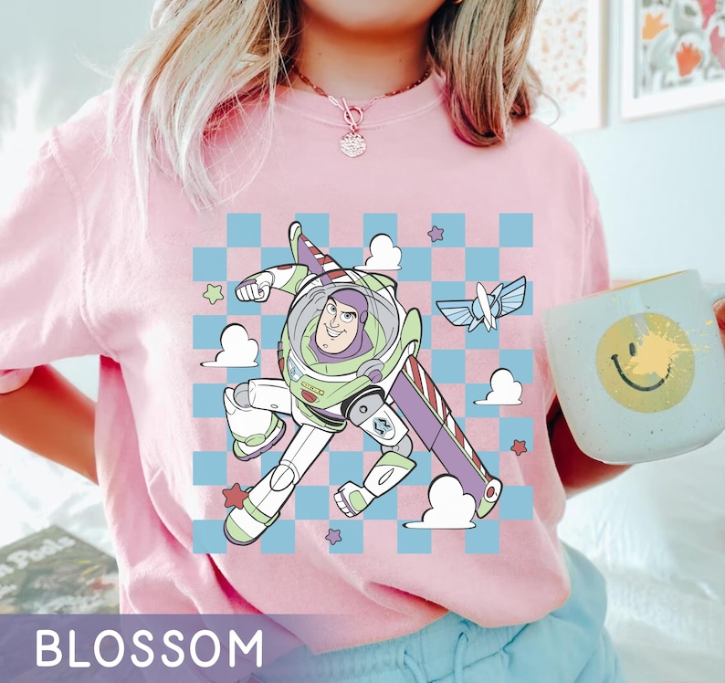 May include: Pink t-shirt with a cartoon image of Buzz Lightyear from Toy Story flying in a spaceship. The image is on a checkered background with clouds and stars.