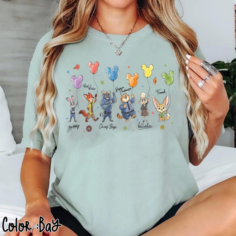 Op de afbeelding: Een lichtgroen t-shirt met stripfiguren, elk met een Mickey Mouse-vormige ballon. De personages zijn onder meer een konijn, een vos, een tijger en anderen. De namen van de personages staan eronder geschreven.