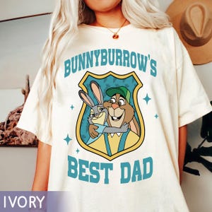 Puede incluir: Camiseta de color marfil con un dibujo animado de dos conejos abraz&aacute;ndose dentro de un escudo. El texto "BUNNYBURROW'S BEST DAD" est&aacute; impreso en turquesa encima y debajo del gr&aacute;fico. La camiseta tiene mangas cortas.