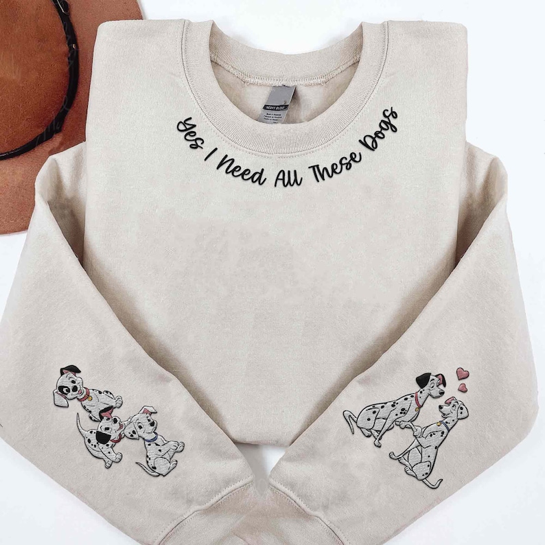 Op de afbeelding: Een beige sweatshirt met de tekst "Yes I need all these dogs" geborduurd rond de halslijn. De mouwen zijn versierd met geborduurde Dalmati&euml;rs.