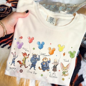 Op de afbeelding: Cr&egrave;mekleurige sweatshirt met stripfiguren uit een film, elk met een Mickey Mouse-vormige ballon. De personages zijn Judy Hopps, Nick Wilde, Chief Bogo, Benjamin Clawhauser, Bellwether en Finnick. De sweatshirt heeft het merklabel Comfort Colors.